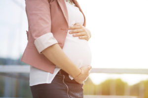 Una futura mamma incinta elegante nella sua giacca rosa accarezza il pancione e sullo sfondo un lago al tramonto